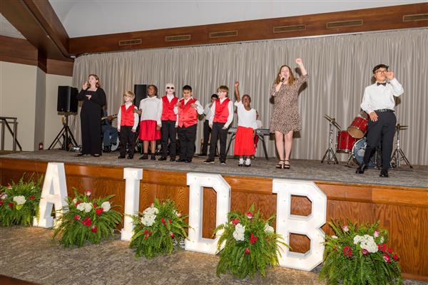 Eight students are lined up on stage wearing coordinated outfits - many in white shirts with red vests or red skirts. Alie B. Gorrie performs with them in a leopard print dress. On the right side, there's a student standing near a drum set. In the foreground at the front of the stage, there are larg